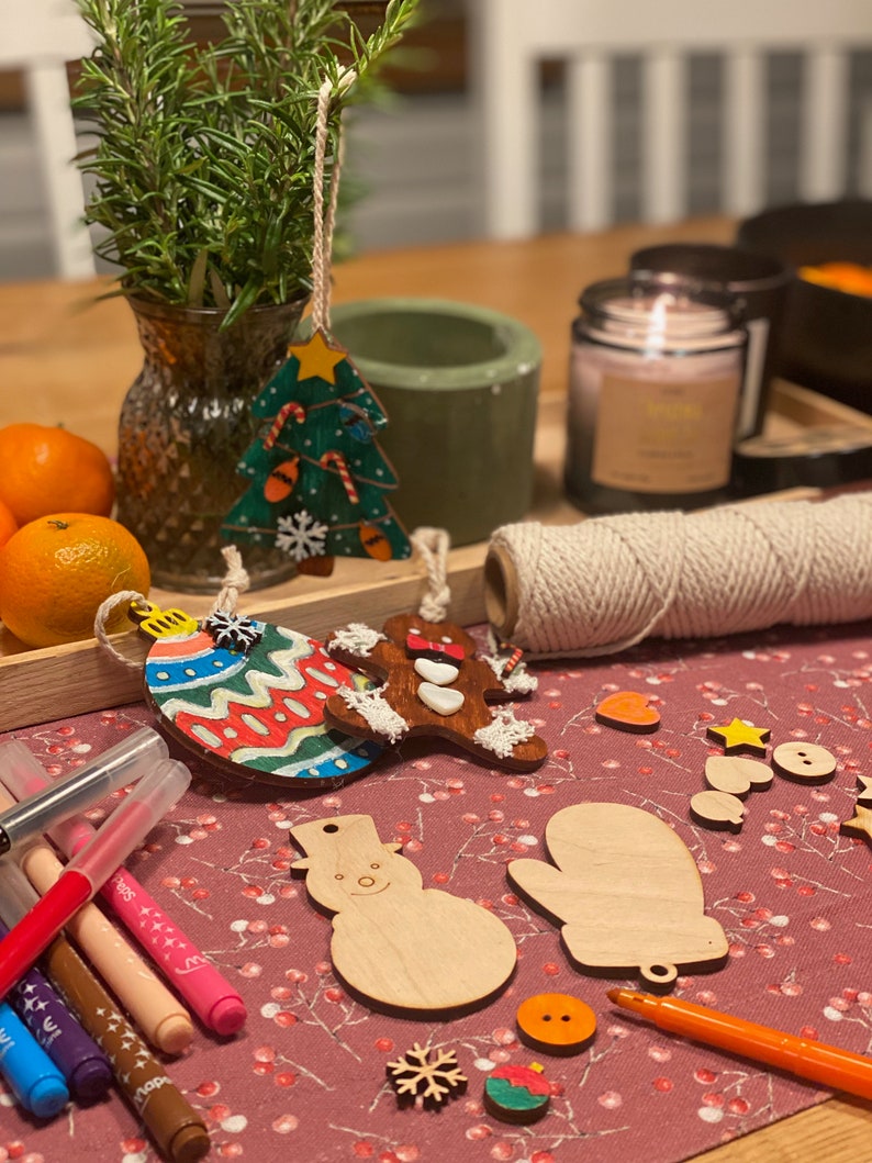 May include: A festive scene with Christmas ornaments, including a tree, gingerbread man, and bauble. Wooden snowman and mitten cutouts are also present, along with markers and twine. The ornaments are on a red patterned surface, suggesting a craft or holiday theme.