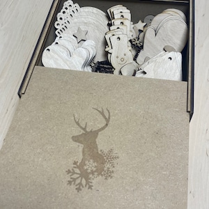 May include: A wooden box with a lid, filled with various laser-cut wooden Christmas ornaments. The ornaments include snowmen, mittens, hearts, stars, and snowflakes. The box lid features a deer silhouette and snowflakes.