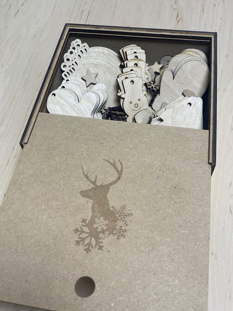May include: A wooden box with a lid, filled with various laser-cut wooden Christmas ornaments. The ornaments include snowmen, mittens, hearts, stars, and snowflakes. The box lid features a deer silhouette and snowflakes.