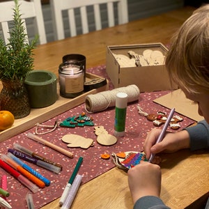 May include: A wooden table with craft supplies, including markers, wooden ornaments, and a candle. A child is coloring a wooden ornament with a purple marker. A small rosemary plant sits in a vase.
