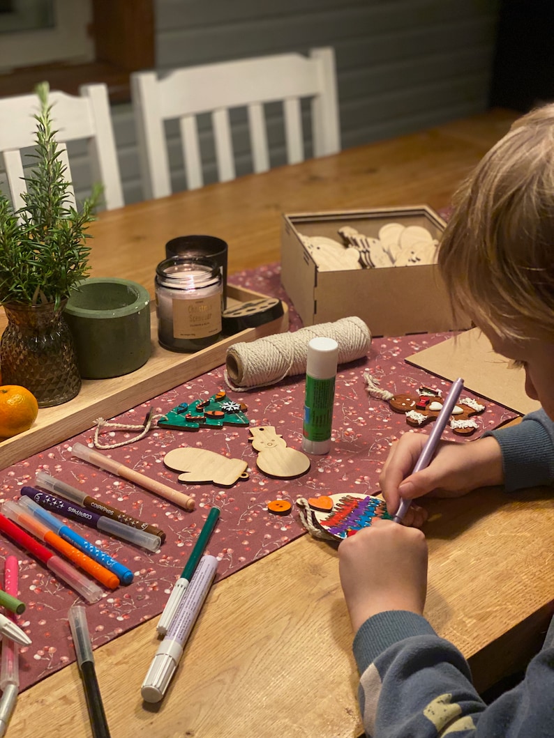 May include: A wooden table with craft supplies, including markers, wooden ornaments, and a candle. A child is coloring a wooden ornament with a purple marker. A small rosemary plant sits in a vase.