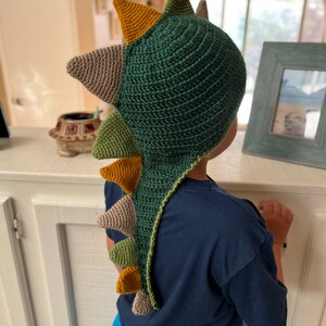 May include: A green, brown, and yellow crocheted dinosaur hat with a long tail. The hat is being worn by a child.
