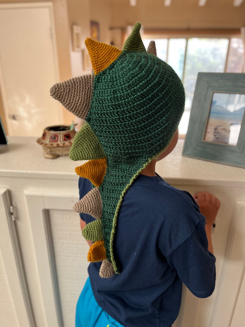 May include: A green, brown, and yellow crocheted dinosaur hat with a long tail. The hat is being worn by a child.