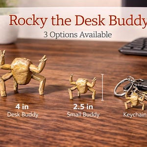May include: The image showcases three gold-colored desk buddies, including a 4-inch desk buddy, a 2.5-inch small buddy, and a keychain version. The text "Rocky the Desk Buddy" and "3 Options Available" is displayed above the items.