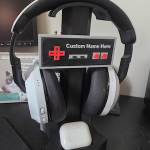 May include: Black headset stand with a retro controller design. The stand holds a white and black headset, with a white case for earbuds at the base. The text "Custom Name Here" is visible on the controller.