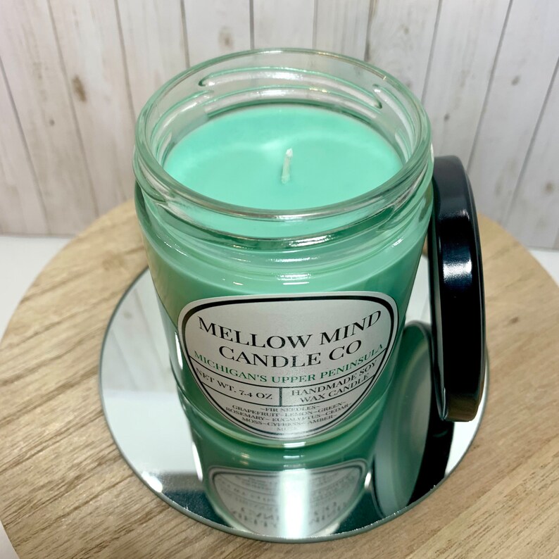 Puede incluir: Una vela de cera de soja verde en un frasco de vidrio con tapa negra. La etiqueta dice "Mellow Mind Candle Co. Michigan's Upper Peninsula. Net Wt. 7.4 oz. Handmade Wax Candle."