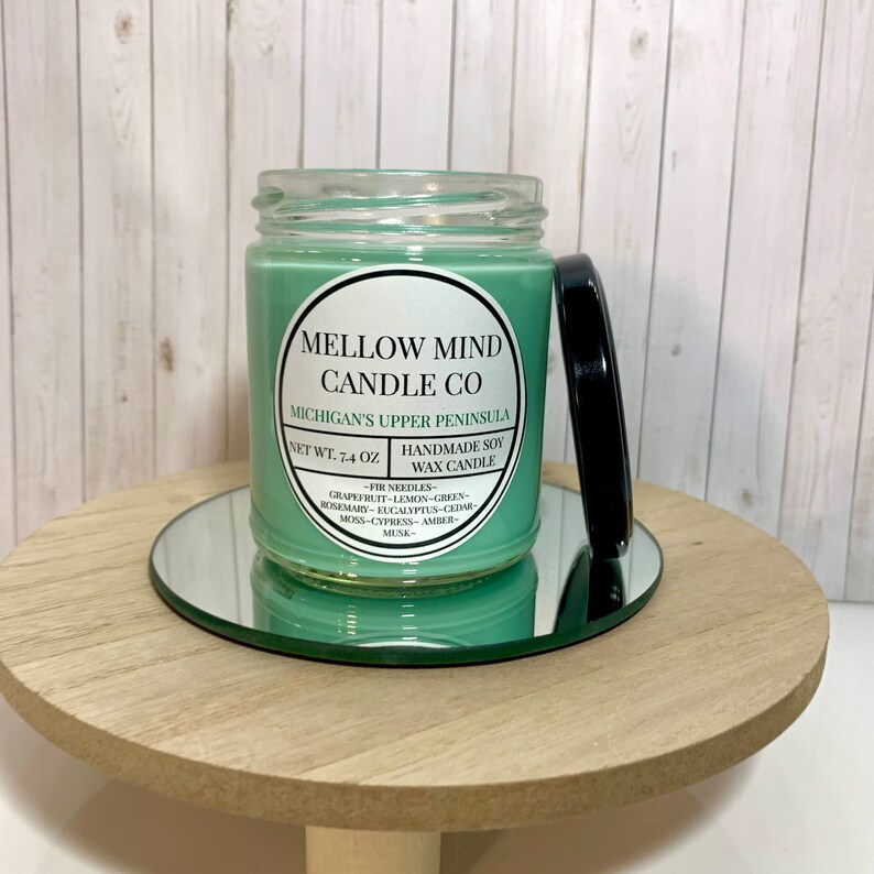 Puede incluir: Una vela de cera de soja verde en un tarro de vidrio con tapa negra. La etiqueta dice "Mellow Mind Candle Co. Michigan's Upper Peninsula. Peso neto: 7,4 oz. Vela de cera de soja hecha a mano - Agujas de pino - Pomelo - Lim&oacute;n - Verde - Romero - Eucalipto - Cedro - Musgo - Cipr&eacute;s - &Aacute;mbar - Almizcle - "