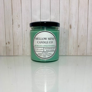 Puede incluir: Una vela de cera de soja verde en un tarro de vidrio con tapa negra. La etiqueta dice "Mellow Mind Candle Co. Michigan's Upper Peninsula. Net Wt. 7.4 oz. Handmade Soy Wax Candle - Fir Needles - Grapefruit - Lemon - Green - Rosemary - Eucalyptus - Cedar - Moss - Cypress - Amber - Musk - "