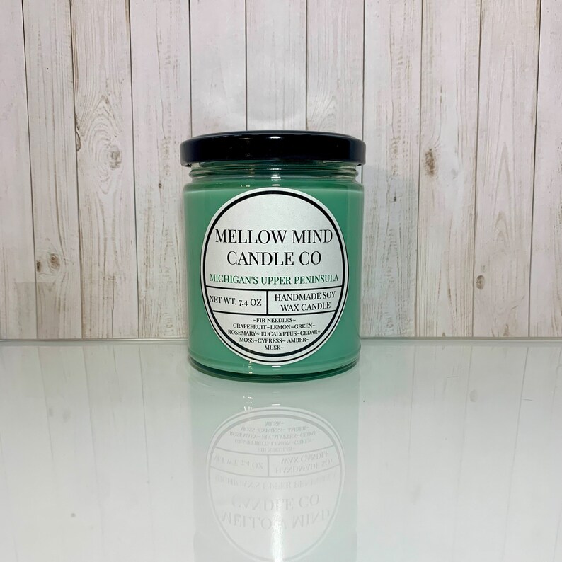 Puede incluir: Una vela de cera de soja verde en un tarro de vidrio con tapa negra. La etiqueta dice "Mellow Mind Candle Co. Michigan's Upper Peninsula. Net Wt. 7.4 oz. Handmade Soy Wax Candle - Fir Needles - Grapefruit - Lemon - Green - Rosemary - Eucalyptus - Cedar - Moss - Cypress - Amber - Musk - "