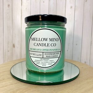 Puede incluir: Un tarro de vela de vidrio verde con una tapa negra. La etiqueta dice "MELLOW MIND CANDLE CO" y "MICHIGAN'S UPPER PENINSULA". La vela es una vela de cera de soja hecha a mano con un peso neto de 7,4 onzas. Las notas de aroma son agujas de pino, pomelo, lim&oacute;n, verde, romero, eucalipto, cedro, musgo, cipr&eacute;s, &aacute;mbar y almizcle.
