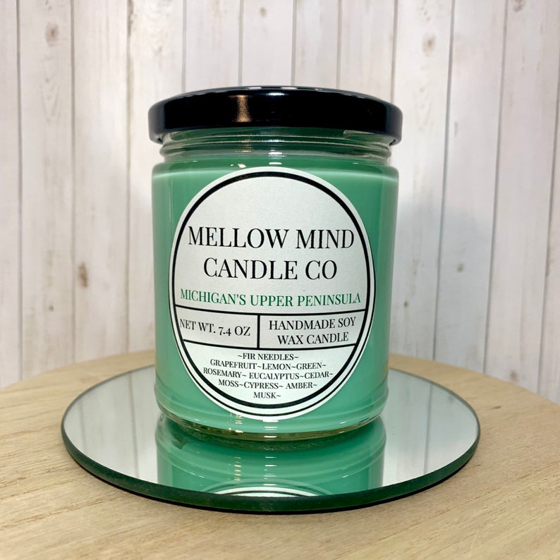 Puede incluir: Un tarro de vela de vidrio verde con una tapa negra. La etiqueta dice "MELLOW MIND CANDLE CO" y "MICHIGAN'S UPPER PENINSULA". La vela es una vela de cera de soja hecha a mano con un peso neto de 7,4 onzas. Las notas de aroma son agujas de pino, pomelo, lim&oacute;n, verde, romero, eucalipto, cedro, musgo, cipr&eacute;s, &aacute;mbar y almizcle.