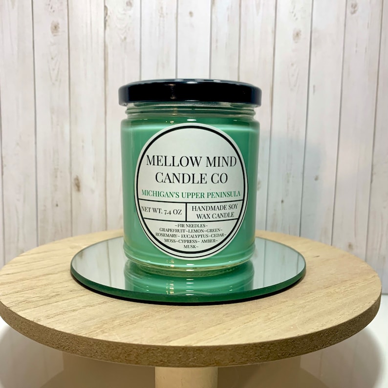 Puede incluir: Un tarro de vela de vidrio verde con una tapa negra. La etiqueta dice "MELLOW MIND CANDLE CO" y "MICHIGAN'S UPPER PENINSULA". La vela es de cera de soja hecha a mano y tiene un peso neto de 7,4 onzas. Las notas de aroma son agujas de pino, pomelo, lim&oacute;n, verde, romero, eucalipto, cedro, musgo, cipr&eacute;s, &aacute;mbar y almizcle.