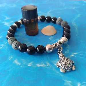 Könnte beinhalten: Schwarzes und graues Perlenarmband mit einem silbernen Schildkrötenanhänger. Das Armband liegt auf einem blauen Hintergrund mit einer kleinen Glasflasche und einer Muschel.