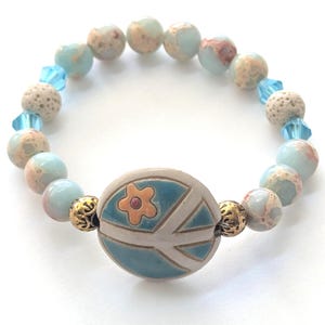 May include: A beaded bracelet featuring a central ceramic bead with a peace sign design and a flower. The bracelet includes light blue, beige, and clear beads. Gold-colored filigree beads accent the central bead.