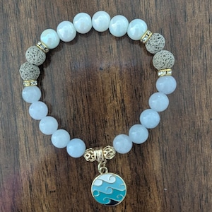 May include: A beaded bracelet featuring light blue and white round beads, with two textured gray beads and gold-colored accents. A round charm with a wave design in shades of blue and white hangs from the bracelet.