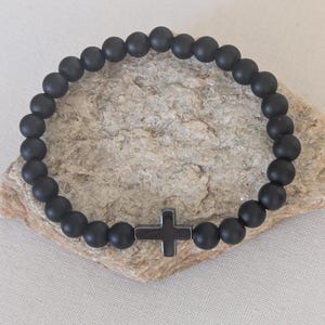 May include: A circular stretch bracelet composed of matte black spherical beads. The bracelet features a single dark gray cross-shaped charm positioned at the bottom center, integrated into the continuous loop of beads.