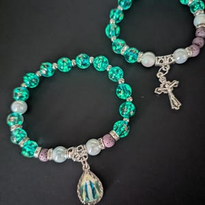 May include: Two beaded bracelets with turquoise and clear beads, white beads, and purple beads. One bracelet has a silver cross charm, and the other has a silver charm with a religious image. The bracelets are on a black background.