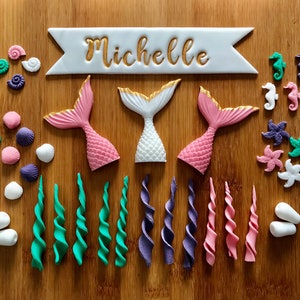 Könnte beinhalten: Ein Set an Kuchendekorationen zum Thema Meerjungfrau, bestehend aus drei rosa und weißen Meerjungfrauen-Schwänzen, einem Banner mit dem Namen "Michelle", Seepferdchen- und Muschelformen sowie grünen und violetten Spiralverzierungen.