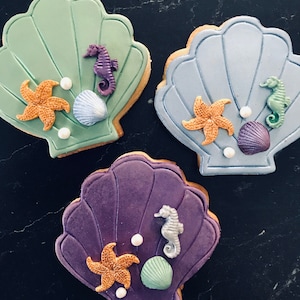 Könnte beinhalten: Drei muschelförmige Kekse mit Dekorationen zum Thema Meer. Jeder Keks hat eine andere Farbe: Grün, Blau und Lila. Sie sind mit Seesternen, Seepferdchen und Muschelbombon verziert, zusammen mit weißen Perlenstreuseln.