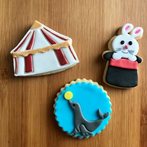 Op de afbeelding: Drie gedecoreerde suikerkoekjes. Een koekje is gevormd als een circustent met rode en gouden strepen. Een ander koekje is gevormd als een konijn dat een witte en rode tovenaarshoed uit een zwarte hoed trekt. Het derde koekje is een blauwe cirkel met een grijze zeehond die een gele bal op zijn neus balanceert.