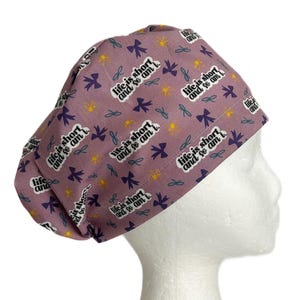 May include: A purple scrub cap with a repeating pattern of bows, flowers, and the text "life is short and so am I." The cap is designed to cover the head and hair, commonly used in medical settings.