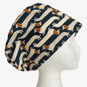 May include: A navy blue and cream-colored patterned scrub cap featuring a repeating design of dachshund dogs. The dogs have brown ears and backs, with cream-colored bodies. The cap is designed to fit over the head and hair.