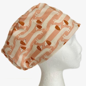 May include: A patterned surgical scrub cap featuring a repeating design of dachshund dogs in a running pose. The cap has a peach and white striped background. The dogs are white with brown ears and backs.