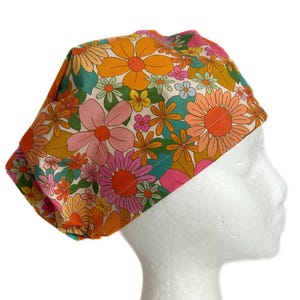 May include: A colorful floral print scrub hat with a white background and a variety of pink, orange, yellow, and green flowers.
