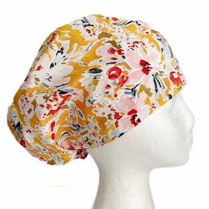 May include: A bouffant-style scrub cap featuring a vibrant floral pattern with mustard yellow, soft pink, bright red, and slate gray brushstroke-style flowers and leaves on a white background, worn on a white mannequin head.