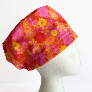 May include: A floral patterned scrub cap in shades of pink, orange, and yellow. The cap is made of fabric and features a repeating pattern of stylized flowers. The cap is shown on a white mannequin head.