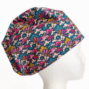 May include: A scrub cap featuring a dense, repeating pattern of stylized clouds and sunbursts in shades of dark teal, magenta, light pink, and mustard yellow. The fabric design includes small teal stars scattered throughout the colorful, layered graphic print.