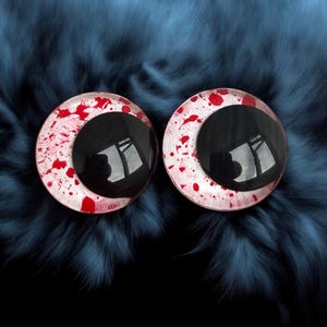May include: A pair of round plastic eyes featuring black pupils and white irises splattered with red. The eyes are set against a dark blue, furry backdrop, creating a stark contrast. The pupils reflect a distorted image.