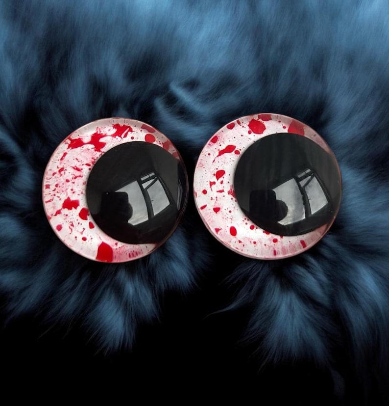 May include: A pair of round plastic eyes featuring black pupils and white irises splattered with red. The eyes are set against a dark blue, furry backdrop, creating a stark contrast. The pupils reflect a distorted image.