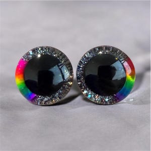 Puede incluir: Dos ojos redondos para manualidades, con centro negro y degradado arcoíris en el borde. Los ojos están rodeados por un borde transparente y brillante. Fondo gris suave.