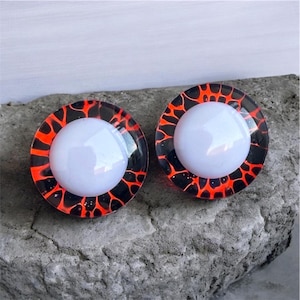 Może przedstawiać: Dwie okrągłe zatyczki, z białym środkiem i czarno-czerwonym wzorem przypominającym lawę na krawędziach. Zatyczki są umieszczone na szarej, teksturowanej powierzchni. Prawdopodobnie są to kolczyki.