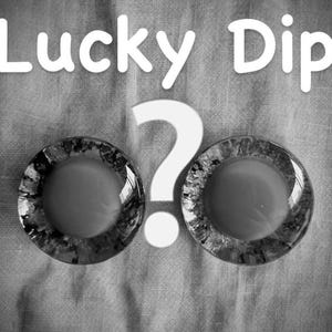 Könnte beinhalten: Schwarz-Weiß-Bild mit dem Text "Lucky Dip" über einem großen weißen Fragezeichen. Zwei runde, transparente Objekte mit strukturiertem Rand und hellem Zentrum sind darunter positioniert.