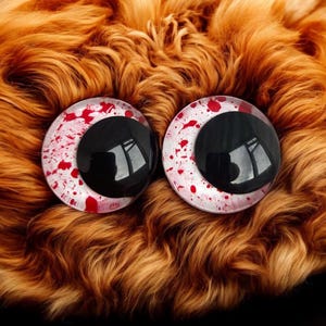 May include: A pair of large, round plastic eyes featuring black pupils and white irises speckled with red. They are set against a backdrop of orange-brown faux fur. These eyes are likely designed for crafting or doll making, offering a dramatic visual effect.