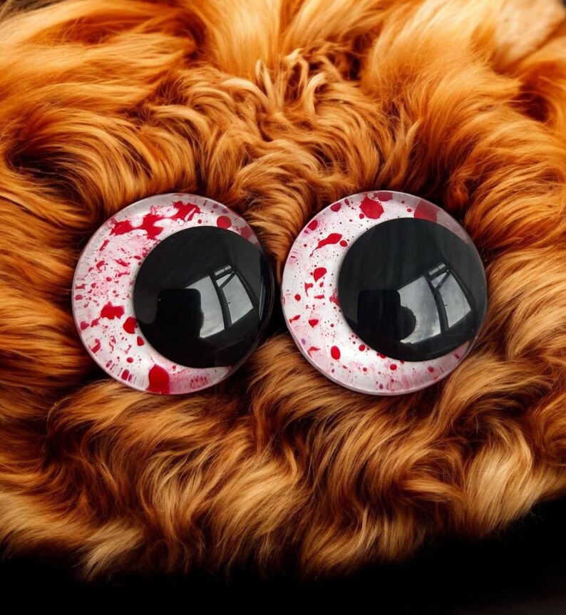 May include: A pair of large, round plastic eyes featuring black pupils and white irises speckled with red. They are set against a backdrop of orange-brown faux fur. These eyes are likely designed for crafting or doll making, offering a dramatic visual effect.