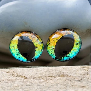 May include: Two circular glass cabochons featuring a realistic animal eye design. The irises display a vibrant gradient of yellow, orange, and teal colours with a textured appearance. Each piece has a large, vertical, black, oval-shaped pupil.