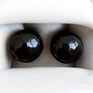 May include: Two large, round, black plugs with a glossy finish, presented in a white, fabric-lined case. The plugs reflect the surrounding environment, creating a mirrored effect. These are likely ear stretching plugs.