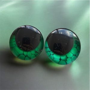 May include: Two circular glass cabochons featuring a realistic reptile eye design. Each piece has a large, solid black pupil centered within a vibrant, translucent green iris that displays a detailed, dark, scale-like honeycomb pattern.