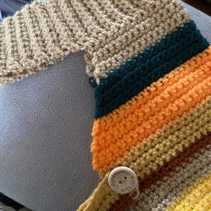 May include: A handmade crochet item featuring a tan sleeve and a striped section in shades of green, orange, yellow, and brown. A wooden button with the text "handmade with love" secures a flap. The item is likely a cozy accessory.