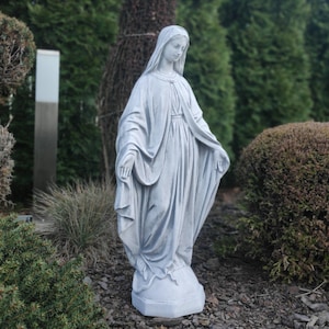Puede incluir: Una estatua gris de una mujer con túnicas fluidas, con las manos abiertas en un gesto de bienvenida. La estatua está situada al aire libre, rodeada de vegetación, y se alza sobre un pedestal.