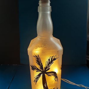 May include: A frosted glass bottle with a cork top, painted with a black silhouette of a palm tree. The bottle is lit from within, creating a warm glow.