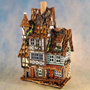 Może przedstawiać: Szczegółowa ceramiczna figurka domu z brązowym, teksturowanym dachem i wieloma piętrami. Fasada posiada okna z białymi ramami, zielone okiennice i czerwone drzwi. Dom jest ozdobiony zielonymi akcentami liści.
