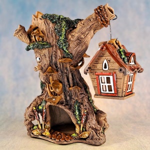 Op de afbeelding: Een keramische boomstam met een klein huis dat eraan hangt. De boomstam is versierd met paddenstoelen en mos. Het huis is rood en bruin met witte ramen.