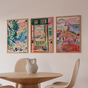 Könnte beinhalten: Drei gerahmte abstrakte Gemälde in einem modernen Esszimmer. Die Gemälde sind in einem farbenfrohen Stil mit einem Mix aus Rosa, Blau, Grün und Gelb gehalten. Die Gemälde hängen an einer weißen Wand über einem Holztisch mit vier weißen Stühlen.