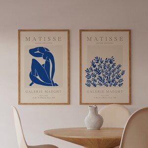 May include: Two framed prints of Matisse artwork, featuring a blue figure and a blue floral design. The prints are titled "MATISSE" and "PAPIER DECOUPES" and are displayed on a white wall above a wooden table with two white chairs.