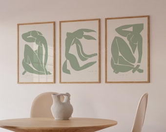 May include: Three framed prints of abstract green figures in various poses, hanging on a white wall above a wooden table with two beige chairs.