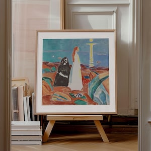 May include: Framed art print depicting a coastal scene with figures. The artwork shows two figures on a beach with a sunset over the sea. The frame is light wood, and the print is displayed on a wooden easel.